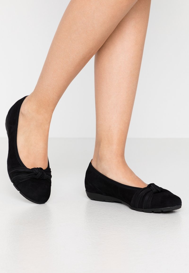 Jambes nues portant des ballerines en daim noir avec un petit détail nœud sur un sol lisse gris clair.
