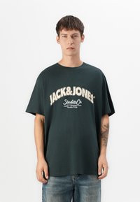 Ung mand med kort brunt hår iklædt en mørk oversized Jack & Jones T-shirt og blå jeans, stående foran en hvid baggrund.