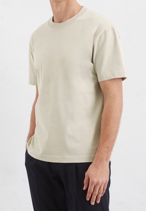 Man draagt een effen beige T-shirt met korte mouwen en ronde hals en donkere broek, staat met één hand in de zak tegen een witte muur.