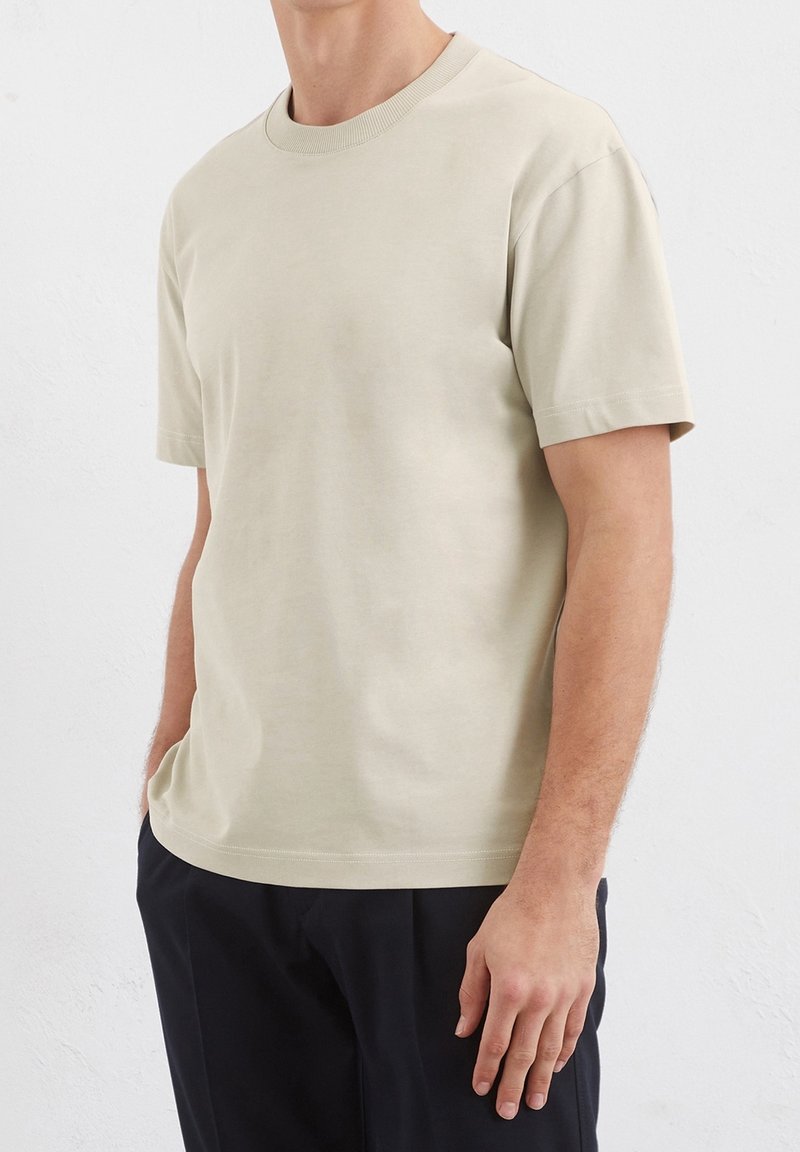 Homme portant un t-shirt beige uni à manches courtes et un pantalon foncé, debout avec une main dans la poche contre un mur blanc.