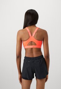 Mujer con cabello largo y oscuro, vistiendo un sujetador deportivo color coral con espalda de nadadora y pantalones cortos deportivos negros, de espaldas contra un fondo claro y sencillo.