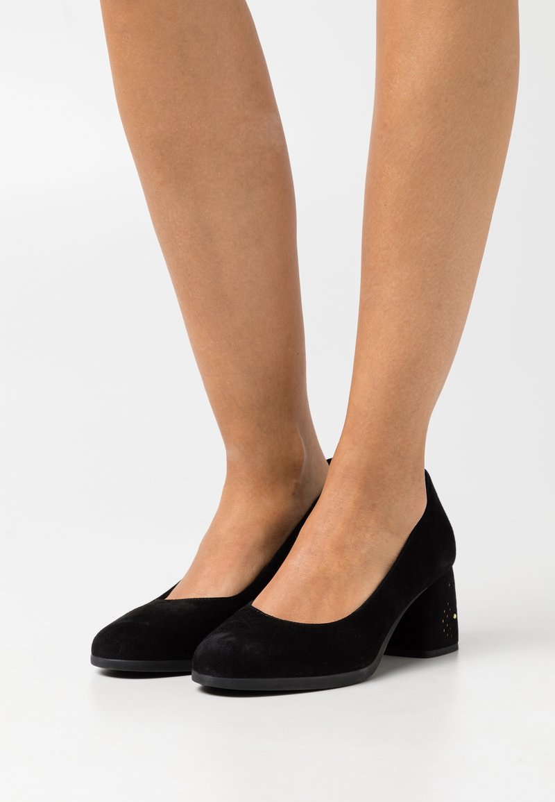 Pieds portant des chaussures à talons blocs noirs en suède à bout fermé avec de subtils éclats dorés sur le talon, posés sur un fond blanc.