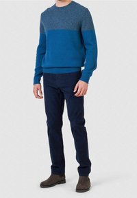 Maglione blu lavorato a maglia con dettagli grigi sulle spalle, collo tondo e polsini a coste, abbinato a pantaloni blu scuro e stivali marroni.