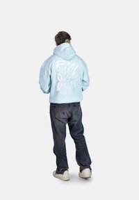Hoodie azzurro chiaro con design in rilievo bianco "Rise & Shine" sul retro, abbinato a jeans scuri e sneakers bianche. Texture liscia, vestibilità casual.