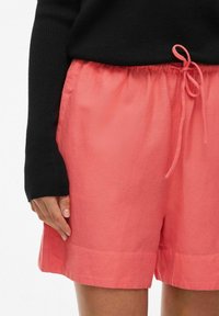 Koralfarvede linnedshorts med elastisk talje og snøre, der har en glat tekstur og foldet kant. Båret med en sort langærmet top.