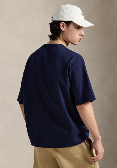 Μπλε ναυτικό βαμβακερό Τ-shirt με κοντά μανίκια και χαλαρή εφαρμογή, συνδυασμένο με μπεζ παντελόνια και λευκό καπέλο. Απλό σχέδιο, χωρίς ορατά μοτίβα.