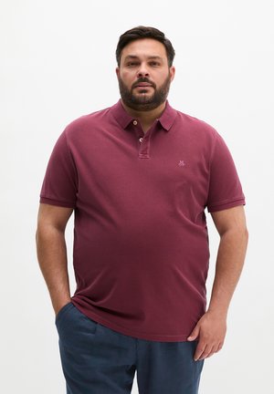 Mężczyzna z brodą, ubrany w bordową koszulkę polo i granatowe spodnie, stoi z jedną ręką w kieszeni na białym tle.