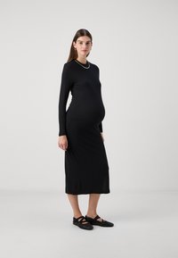 Μαύρο ριμπ φόρεμα εγκυμοσύνης με μακριά μανίκια, εφαρμοστή γραμμή, μήκος μέχρι τον αστράγαλο και στρογγυλή λαιμόκοψη, συνδυασμένο με μαύρες φλατ πέδιλες.