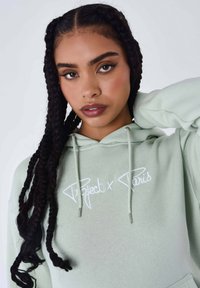 Felpa verde chiaro con testo ricamato "Project x Paris", dotata di coulisse e una texture morbida. Il modello ha lunghi capelli intrecciati.