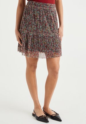 Kvinde iført et plisseret blomsterprintet skørt med flæsekant og sorte spidse slingback-flats, stående mod en ensfarvet baggrund.