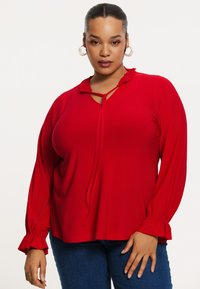 Rød bluse med lange ærmer i blødt materiale, med flæser ved halsudskæringen og manchetterne, der har en nøglehuludskæring og bindebånd foran.