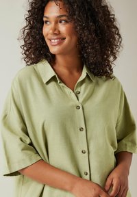 Camicia verde chiaro con bottoni e maniche corte, realizzata in un tessuto testurizzato. Presenta un colletto classico e bottoni marroni lungo la parte anteriore.