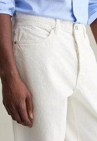 Los jeans de mezclilla blanca tienen un corte recto, bolsillos estándar y un cierre de botón metálico. La tela tiene una textura suave con costuras sutiles.