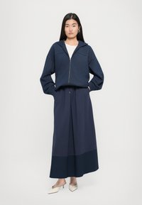 Dunkelblauer Zip-Hoodie mit Kapuze und elastischem Bund, kombiniert mit einem langen, fließenden dunkelblauen Rock mit Farbblockdesign. Beigefarbene Mules runden den Look ab.