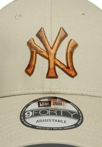 Beige justerbar caps med et brodert brunt New York Yankees-logo. Glatt stoff med en teksturert overflate, rund skygge og innvendig etikett.