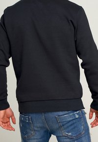 Felpa nera con un design semplice, polsini e orlo a costine, indossata sopra jeans blu strappati. Vista posteriore che mostra la texture del tessuto e la vestibilità.
