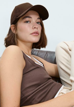 Hnedý rebrovaný top bez rukávov nosený pod bielou košou, doplnený tmavohnedou čiapkou. Osoba má krátke vlasy a neutrálny pozadie.