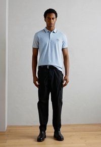 Camisa polo azul claro con cuello y ribete negro contrastante, combinada con pantalones negros y botas negras, de pie sobre un suelo de madera.