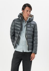 Gray puffer jacket featuring a hood, quilted design, front zipper, and pockets. Worn over a light blue shirt with dark jeans.