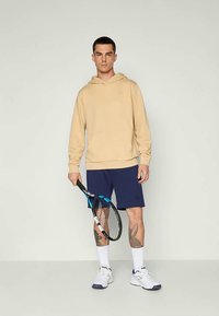 Bézs krāsas kapuču džemperis ar priekšējo kabatu, tumši zili šorti, baltas zeķes un sporta apavi, turot tenisa raketi.