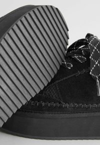 Zapato de ante negro con suela de goma gruesa y estriada, acentos texturizados y cordones con patrón. La vista en primer plano enfatiza el material y el diseño.