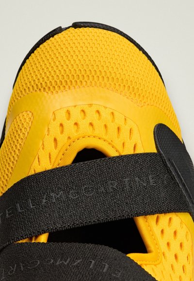 Zapatilla deportiva amarilla con parte superior de malla transpirable, suela de goma y una ancha tira negra a lo largo del mediopié con texto en relieve.