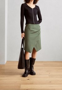 Maglia nera a costine con maniche lunghe e zip frontale, abbinata a una gonna in jersey verde con orlo asimmetrico. Stivali marroni al ginocchio e una borsa tote nera.