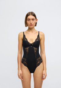 Μαύρο κορμάκι με δαντελένιες λεπτομέρειες και ρυθμιζόμενους ιμάντες, διαθέτει βαθύ ντεκολτέ και εφαρμοστή σιλουέτα. Ομαλή υφή με περίπλοκα σχέδια.