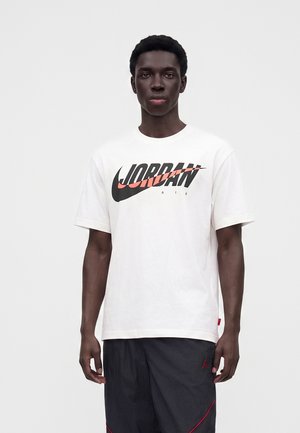 Jeune homme portant un t-shirt blanc Jordan Air avec un logo noir et orange et un short noir avec des détails rouges, debout devant un fond uni.