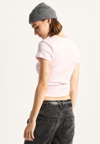 Hellrosa gerippter Kurzarm-Crop-Top, kombiniert mit hochgeschnittenen schwarzen Jeans und einem strukturierten schwarzen Gürtel, von hinten dargestellt.