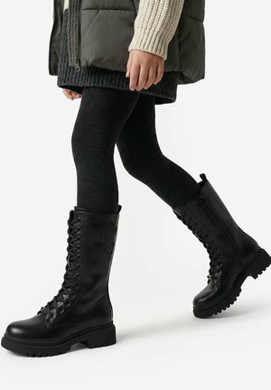Kind trägt schwarze kniehohe Stiefel mit Herzpatches, schwarze Strumpfhosen, einen Strickrock und eine Steppjacke und läuft vor weißem Hintergrund.
