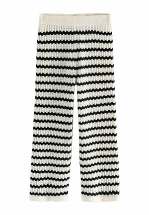 Nohy obuté v béžových semišových topánkach na pracku a čierno-bielo prúžkovaných pletených nohaviciach, zobrazené na neutrálnom pozadí.