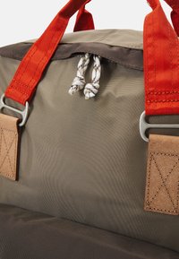 Canvas duffel bag with orange webbing handles, brown zipper, and beige body. Features leather accents and knotted drawstring closure.