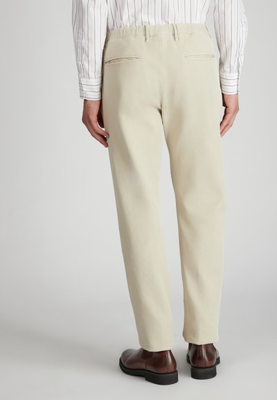 Beżowe, dopasowane spodnie o gładkiej teksturze, z dwiema tylnymi kieszeniami i eleganckim krojem, w połączeniu z ciemnobrązowymi butami wizytowymi.