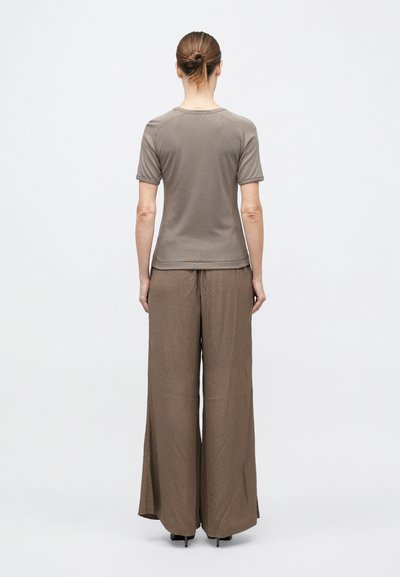 Persona ar matu saišķi, tērpusies īsroku taupe krāsas topā un brūnās platajās biksēs, stāvot ar muguru pret vienkāršu baltu fonu.