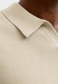 Polo en maille beige avec col plat. La texture est légèrement côtelée, mettant en valeur un design simple et épuré.