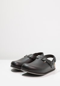 Zoccoli neri con finitura opaca, cinturino regolabile, punta arrotondata e suole testurizzate. Accento in sughero visibile al tallone, progettati per il comfort.