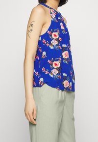 Niebieska bezrękawnikowa bluzka w kwiatowy wzór z plisowanym wykończeniem, wykonana z lekkiego materiału. W połączeniu z jasnobrązowymi spodniami.