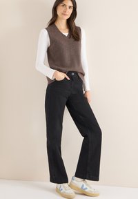 Braune Strickweste über einem weißen Langarmshirt, kombiniert mit hoch taillierten schwarzen Jeans und weißen Sneakern mit blauen Akzenten.