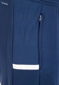 Nahaufnahme eines dunkelblauen Stoffes mit strukturiertem Muster, einer Reißverschlusstasche, einem weißen horizontalen Streifen und "Climacool"-Branding.