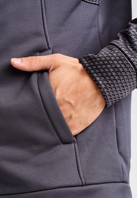 Felpa grigia scura con maniche textured e a motivo. Presenta una tasca laterale con una mano appoggiata all'interno. Tessuto morbido e liscio in vista.