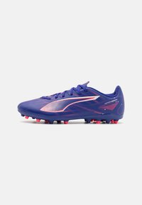 Botín de fútbol morado con una superficie texturizada, detalles en blanco y tachuelas rojas. Presenta un perfil bajo y un diseño con cordones.