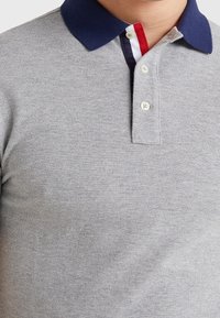 Grå polo shirt med marinblå krage, utrustad med en tre-knappsslutning och kontrasterande röda och vita detaljer inuti kragen. Strukturerad tyg.