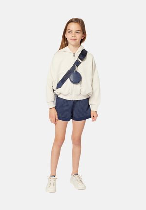 Sweat à capuche zippé blanc, short plissé marine, baskets blanches et sac en bandoulière rond marine avec une sangle. Fond neutre, pose debout.