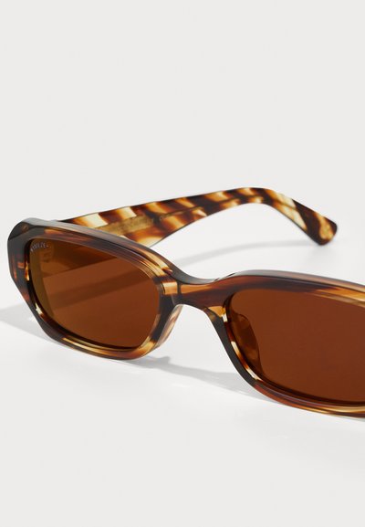 Lunettes de soleil écaille de tortue brunes avec des verres brun foncé. La monture a une finition en plastique lisse et des bords arrondis, avec un design de branche à motifs.