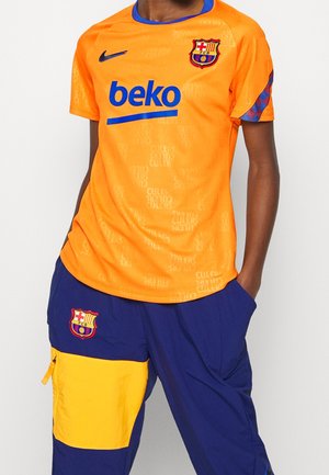 Persoon draagt een oranje FC Barcelona voetbalshirt met blauw "beko" logo en donkerblauwe broek met een gele zak en het FC Barcelona embleem.