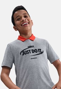Polo gris de Nike con un cuello naranja, que presenta un gran gráfico negro "JUST DO IT" en la parte frontal y un detalle de texto técnico debajo.