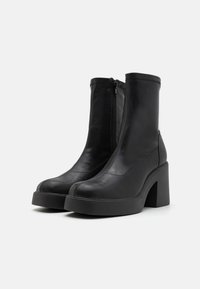 Bottines en cuir noir avec un talon épais, dotées d'une fermeture éclair verticale à l'avant et d'une texture lisse. Design simple avec des accents minimalistes.