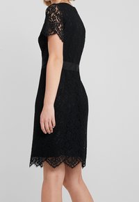 Schwarzes Spitzekleid mit kurzen Ärmeln, ausgestattet mit einem samtigen Taillenbund und einem gewellten Saum. Detailliertes florales Spitzenmuster über das ganze Kleid.