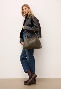 Olíva zöld bőr kézitáska textúrált felülettel, arany színű fém kiegészítőkkel és laza fazonnal. Sötét dzsekivel és kék farmernadrággal viselve.